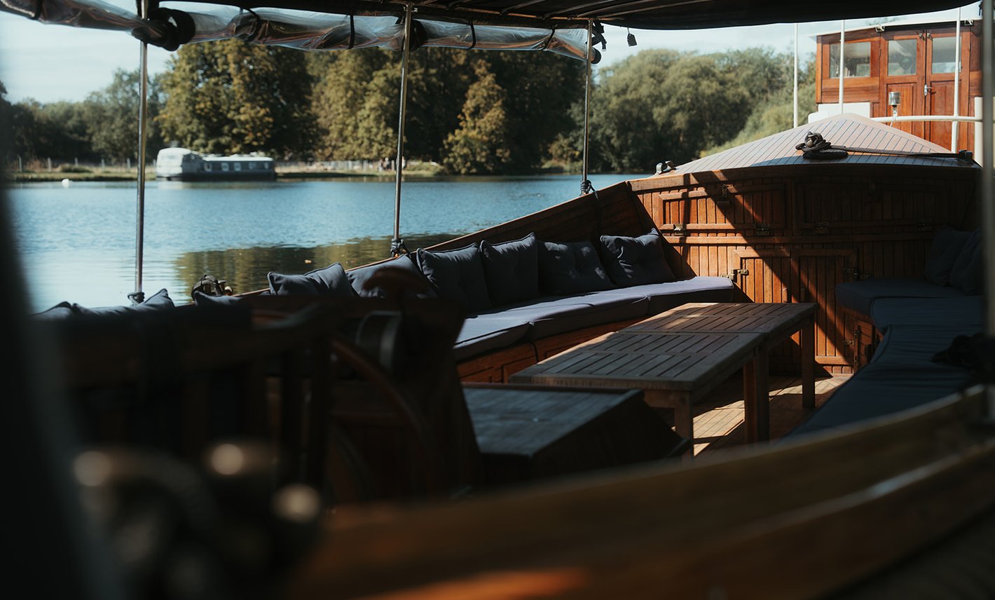 Sunlit cushioned seating and teak tables aboard, looking out across the Thames