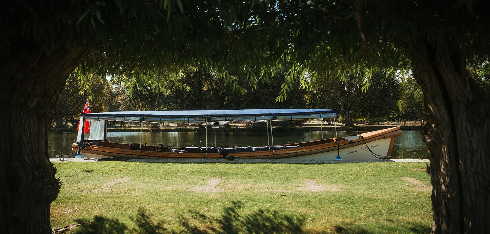 Queen of the Slipstream moored on a sunlit riverbank, framed by willow trees